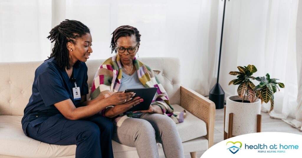 A caregiver talks with a patient while showing her information on a tablet representing the type of thorough communication needed for effective and professional care of home health care patients.