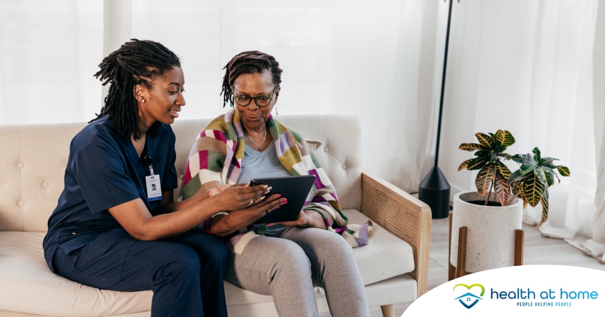 A caregiver talks with a patient while showing her information on a tablet representing the type of thorough communication needed for effective and professional care of home health care patients.