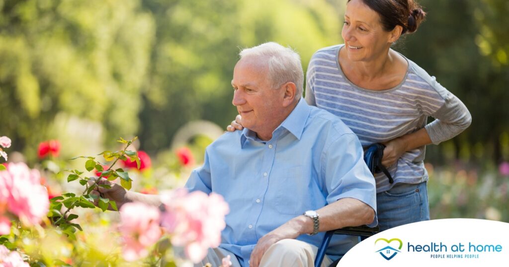A caregiver brings an older adult with a wheelchair to see some flowers representing how professional caregivers can make outdoor activities accessible.