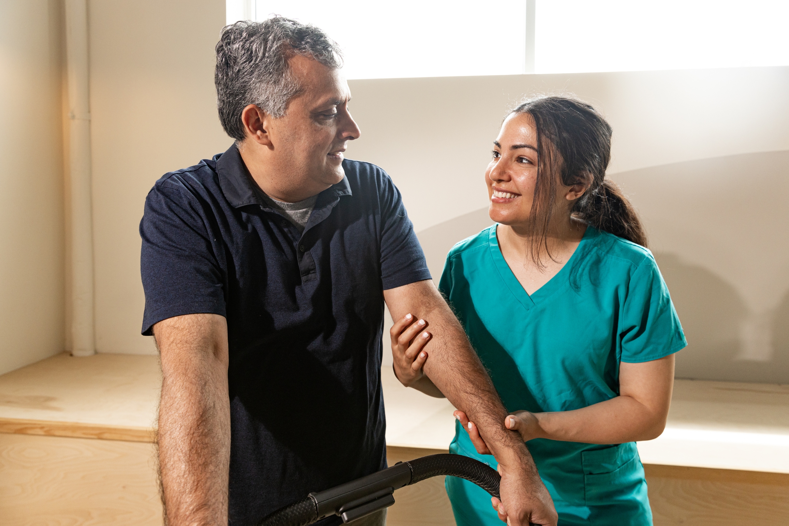 Senior home care in Palm Beach Gardens A smiling female caregiver in medical scrubs gently supports and talks with an elderly man in a bright, relaxed home. This image reflects compassionate senior home care in Palm Beach Gardens, IA, highlighting trust, meaningful connection, and personalized support in a warm, comfortable setting.