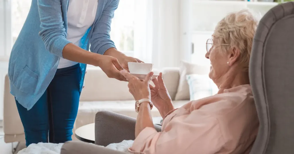 private duty caregiver giving home care client sitting in chair a cup of tea 