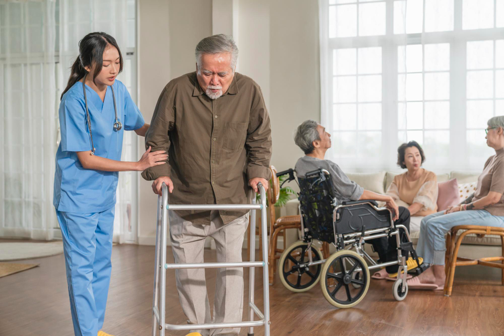 Caregiver in scrubs helping a senior man walk with a walker while family looks on