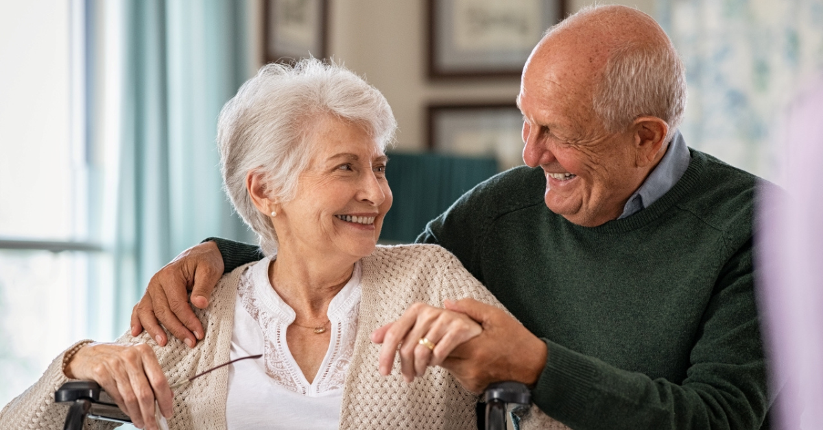 Elderly couple smiling at each other, woman in wheelchair, illustrating couples care