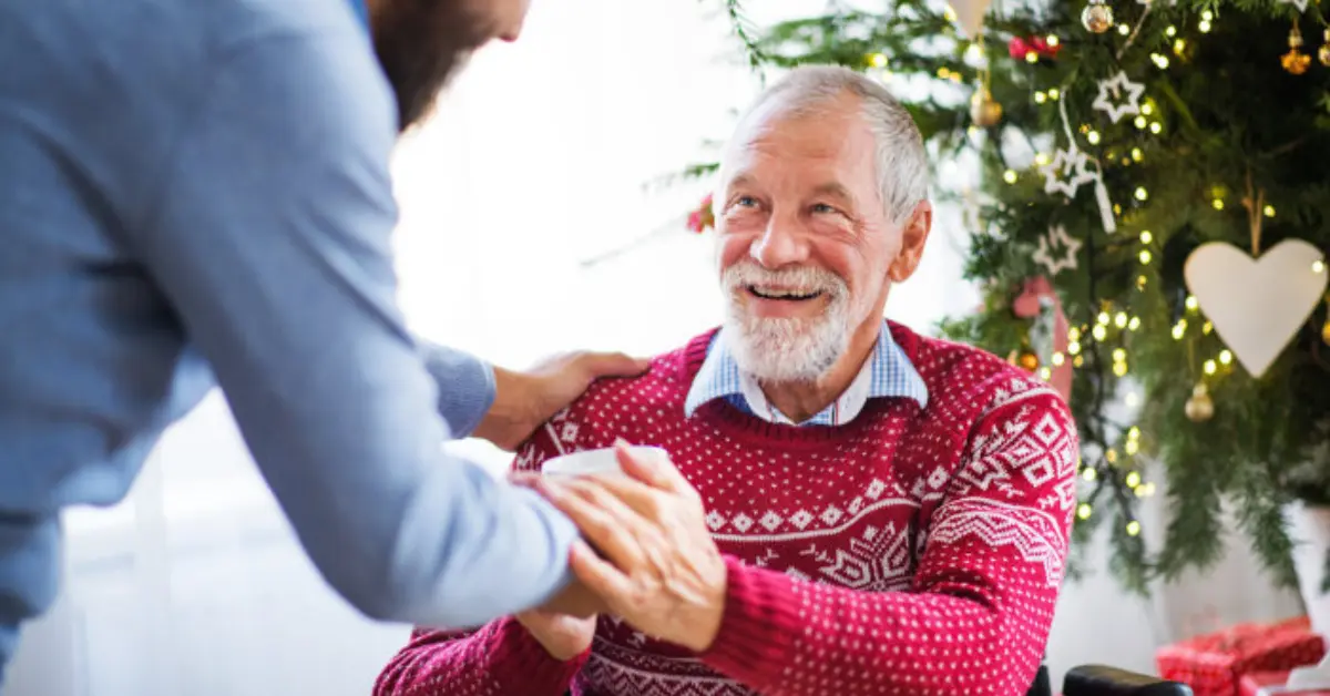 Elderly man in wheelchair receiving a gift from younger man near Christmas tree