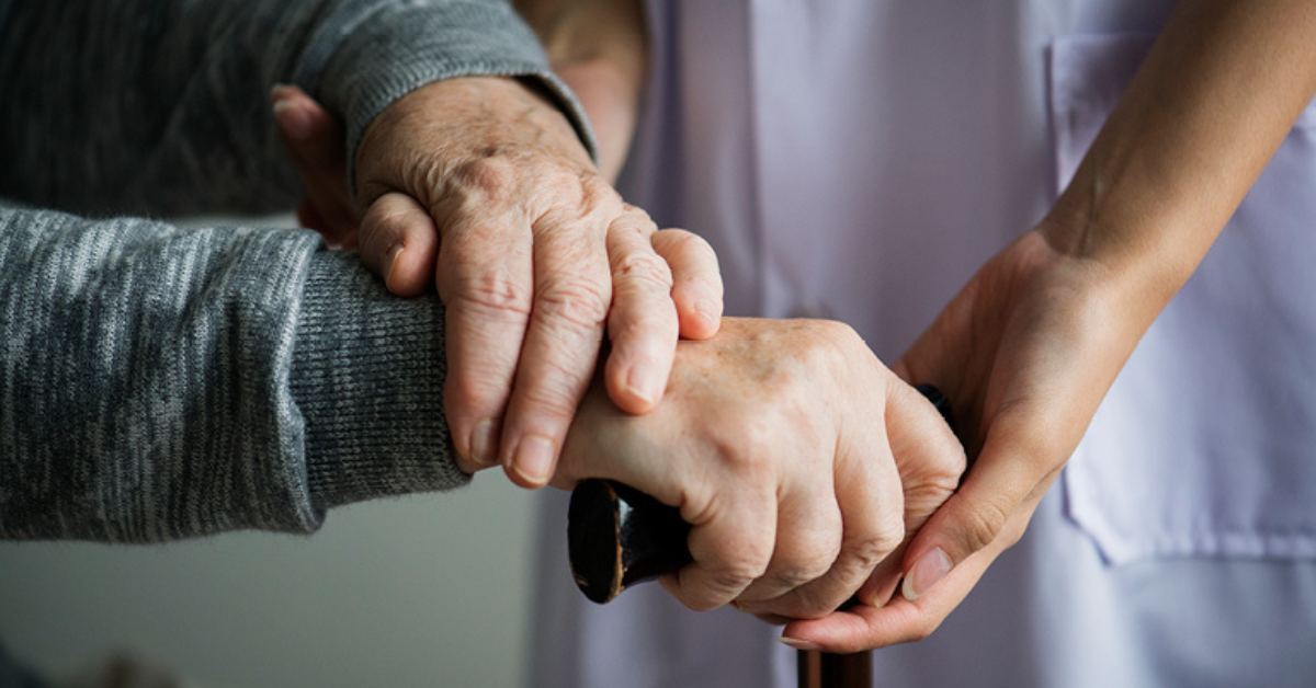 Caregiver's hands supporting an elderly person's hand on a cane, signifying parkinson's care.