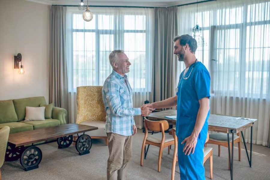 Doctor in blue scrubs shakes hands with a senior man in a living room.
